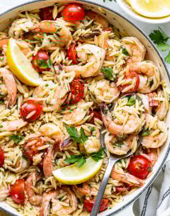 A skillet filled with orzo pasta, shrimp, cherry tomatoes, parsley, and lemon wedges, with a serving spoon resting inside.