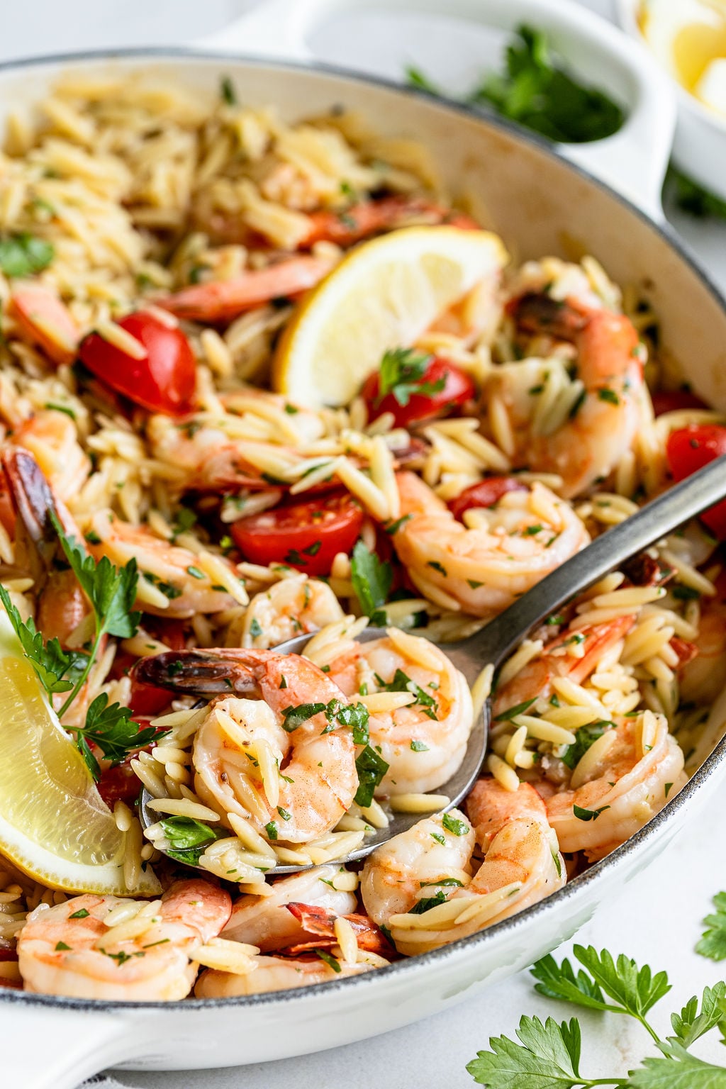 A skillet filled with cooked shrimp, orzo pasta, cherry tomatoes, parsley, and lemon wedges, with a serving spoon scooping some out.