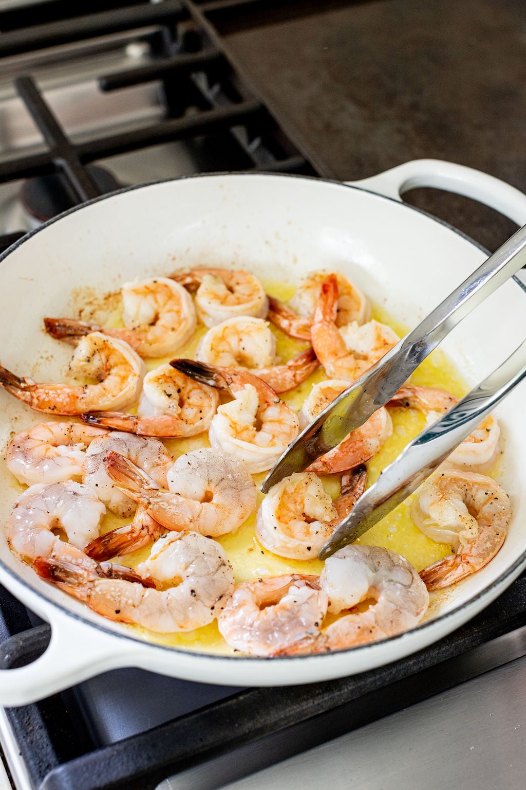 Raw and cooked shrimp being saut&eacute;ed in a white skillet on a stovetop, with metal tongs holding one shrimp.