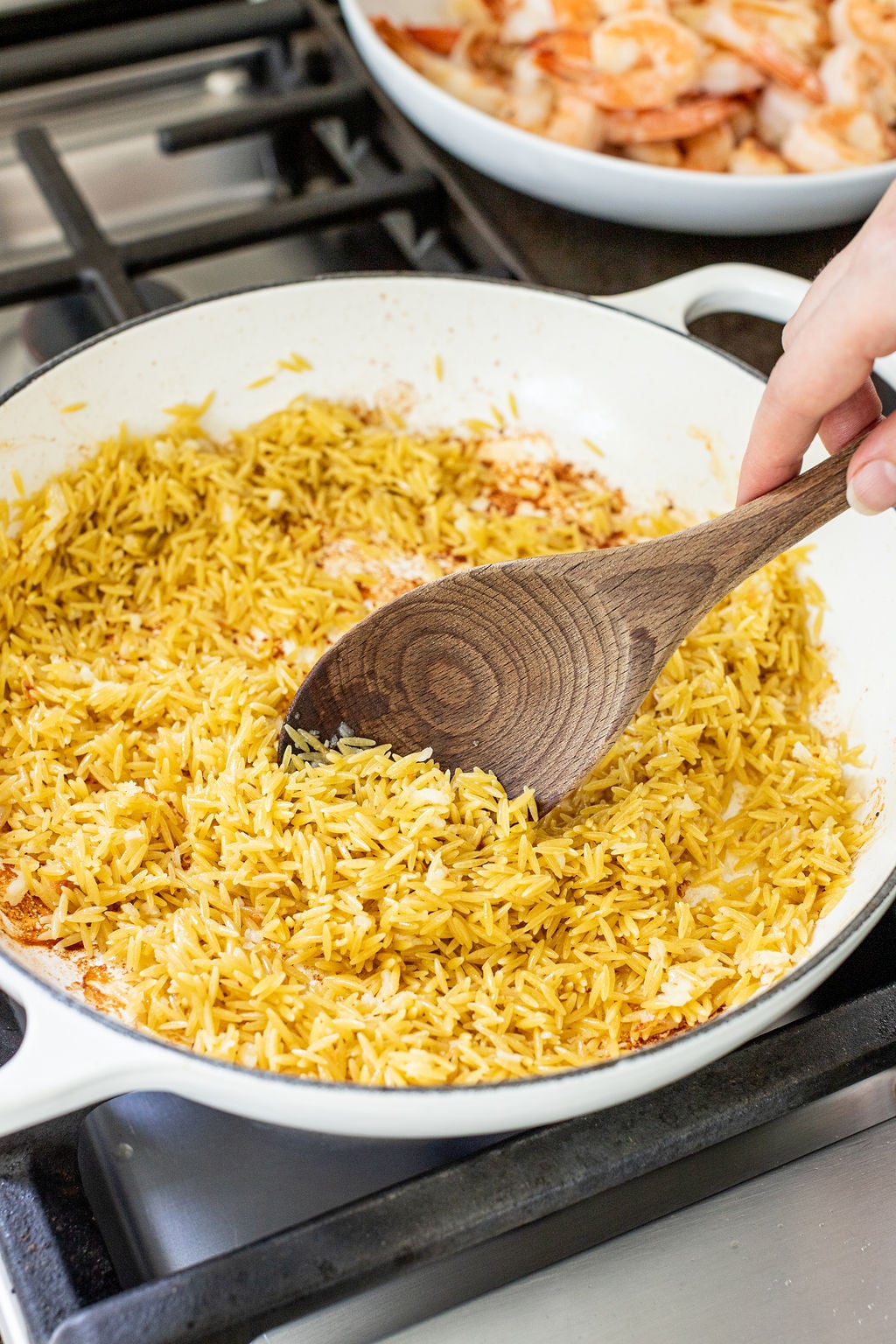A hand stirring yellow rice in a white skillet on a stovetop with a wooden spoon.