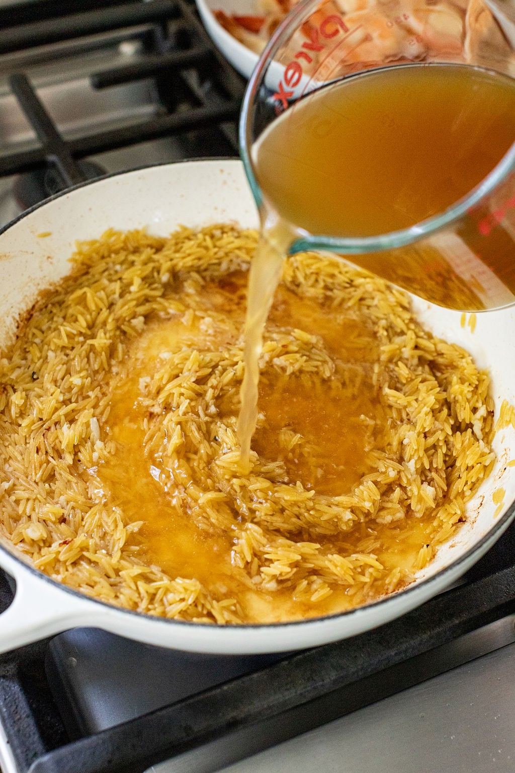 A clear measuring cup pours broth into a skillet with browned orzo pasta on a stovetop.