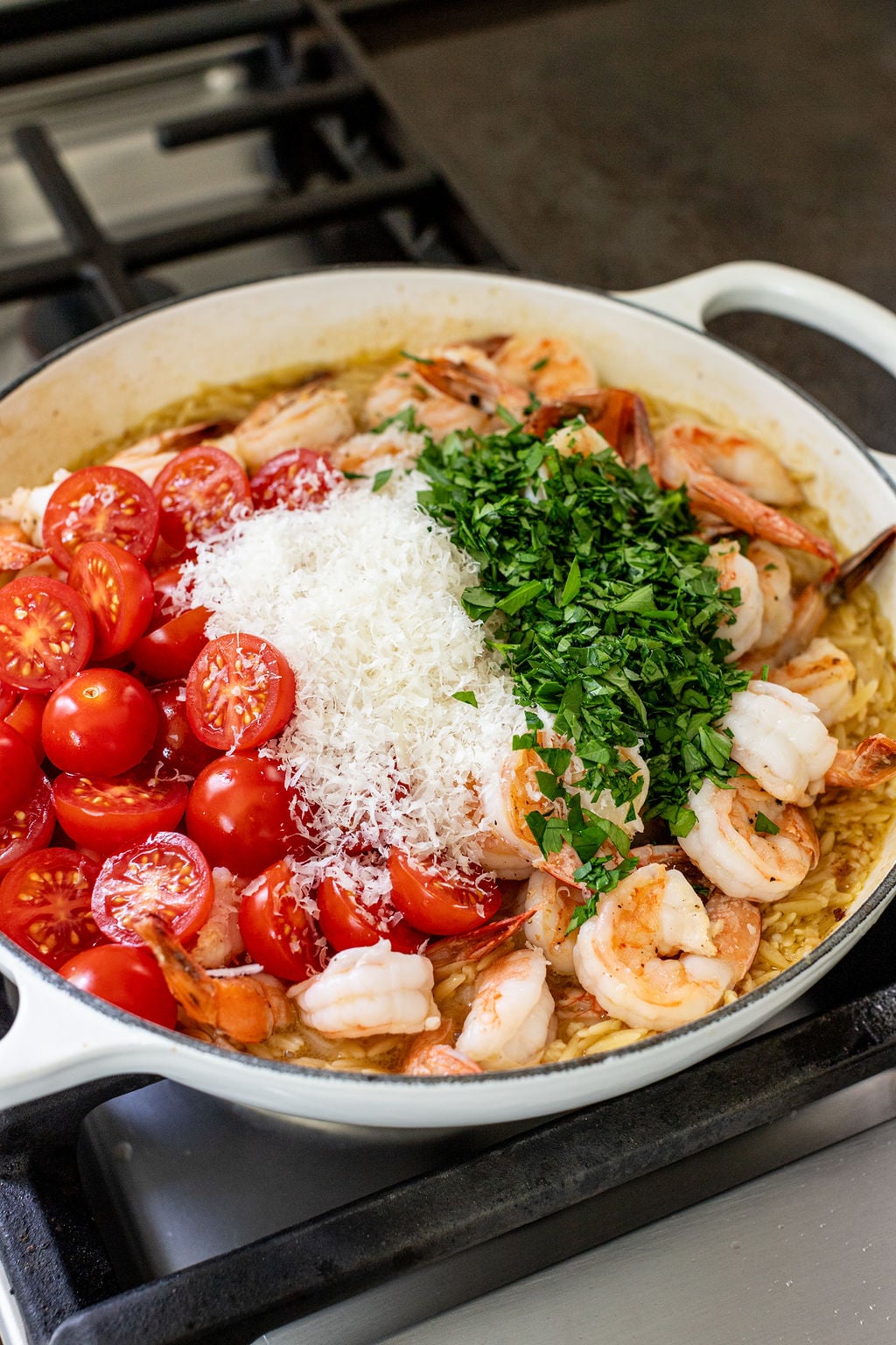 A skillet on a stove contains shrimp, halved cherry tomatoes, grated cheese, and chopped herbs.