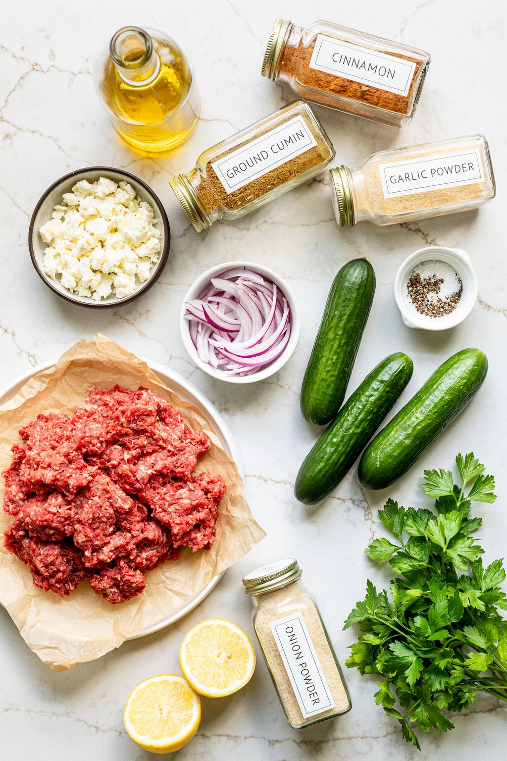 Ground beef, feta cheese, red onion, cucumbers, parsley, lemon halves, olive oil, spices, and black pepper on a marble surface.