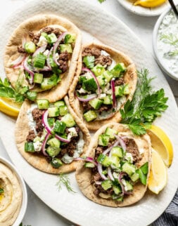 Four pita breads topped with ground meat, cucumber salad, red onion, feta cheese, and fresh herbs are arranged on a white platter with lemon wedges.