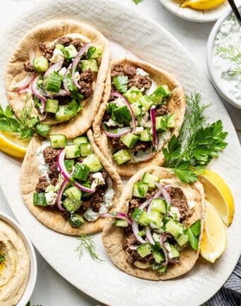 Four pita breads topped with ground meat, cucumber salad, red onion, feta cheese, and fresh herbs are arranged on a white platter with lemon wedges.
