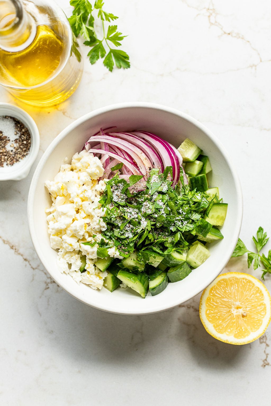 A bowl containing chopped cucumber, red onion, crumbled feta cheese, and fresh herbs, with salt and pepper on top.