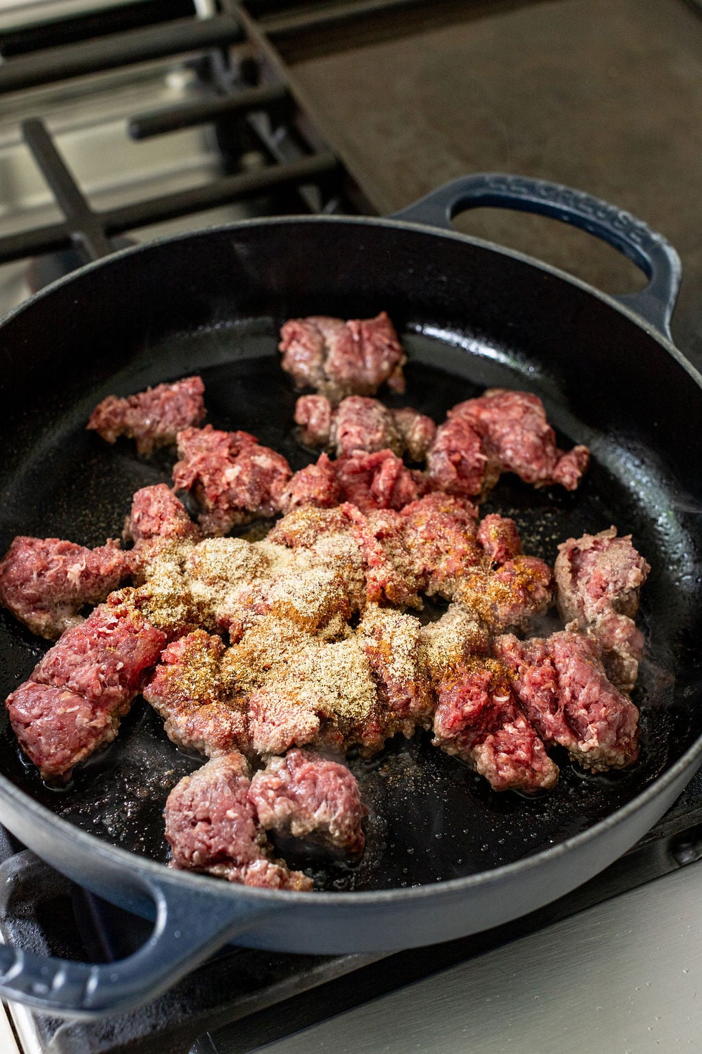 Ground beef with seasonings cooking in a black cast iron skillet on a stovetop.