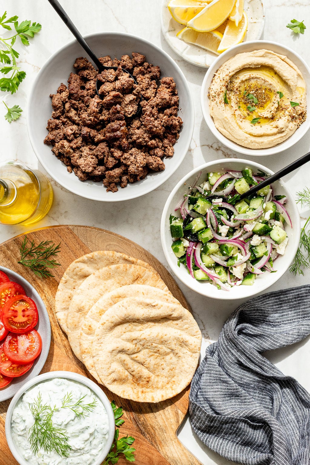 A flat lay of Mediterranean ingredients including cooked ground meat, hummus, cucumber salad, pita bread, sliced tomatoes, tzatziki, lemon wedges, olive oil, and herbs.