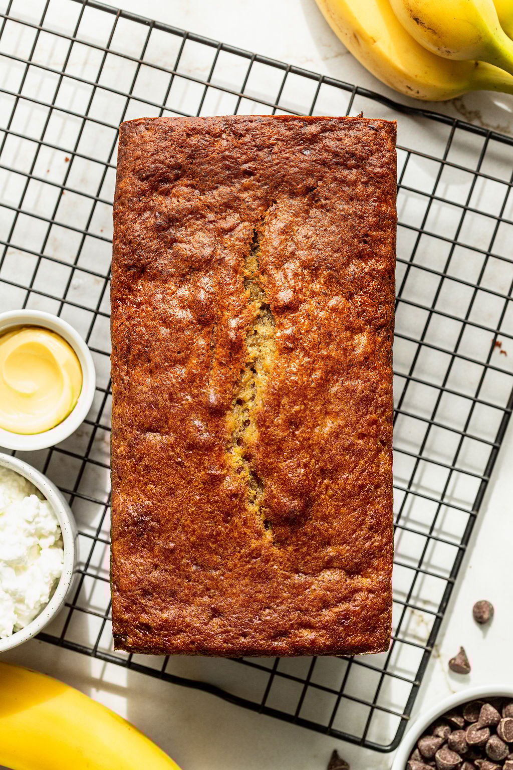 A loaf of banana bread rests on a wire cooling rack, surrounded by bananas, chocolate chips, and a small bowl of butter.
