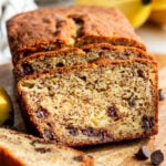 Sliced loaf of banana bread with chocolate chips on a wooden board, with bananas and chocolate chips in the background.