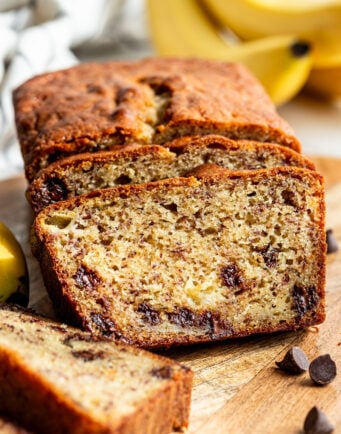 Sliced loaf of banana bread with chocolate chips on a wooden board, with bananas and chocolate chips in the background.