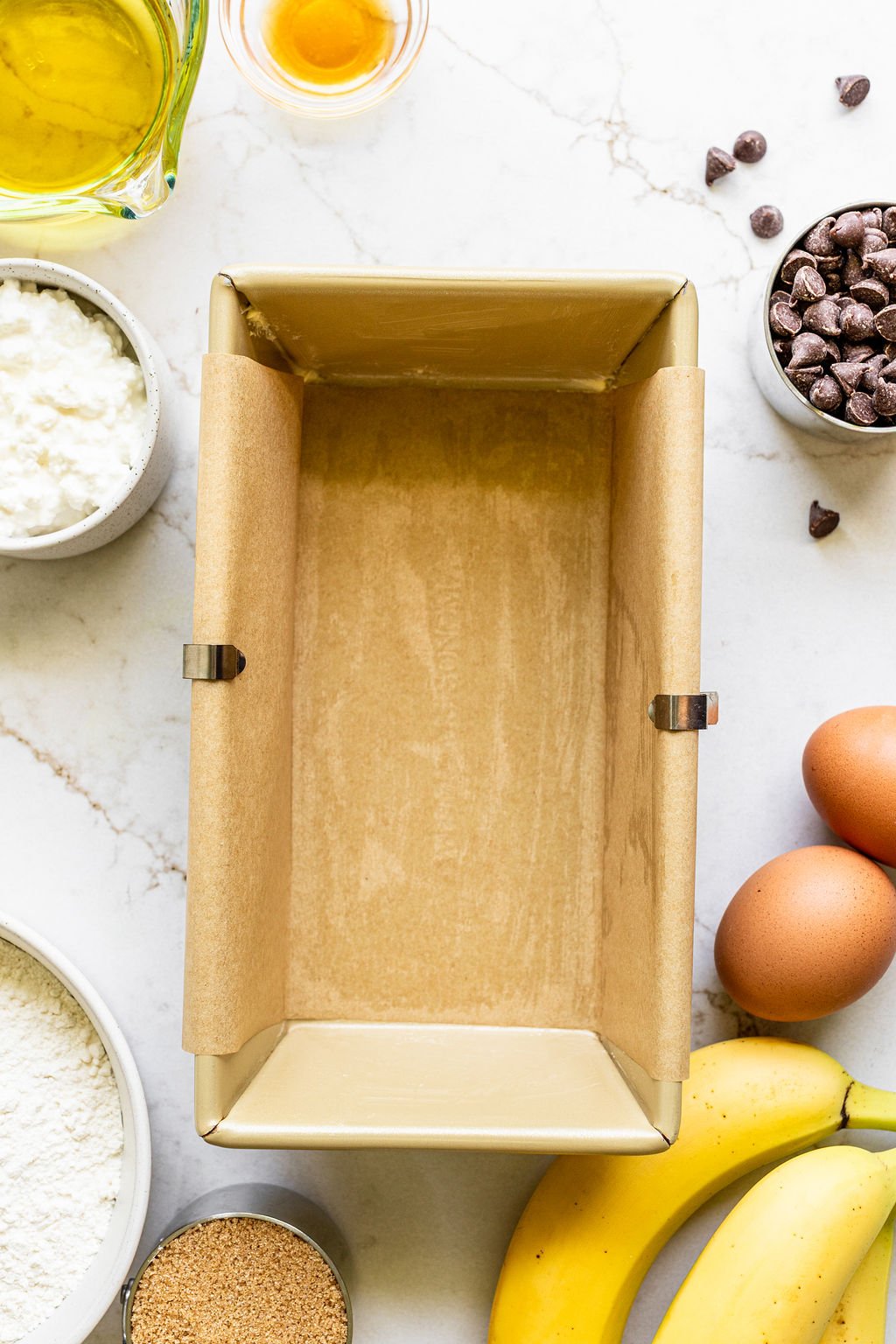 A parchment-lined loaf pan surrounded by baking ingredients including bananas, eggs, chocolate chips, flour, sugar, cottage cheese, oil, and vanilla on a white surface.