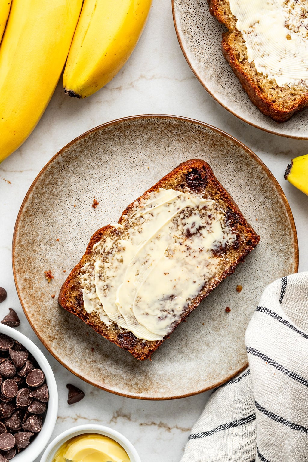A slice of banana bread with butter on a plate, surrounded by bananas, chocolate chips, and a napkin.