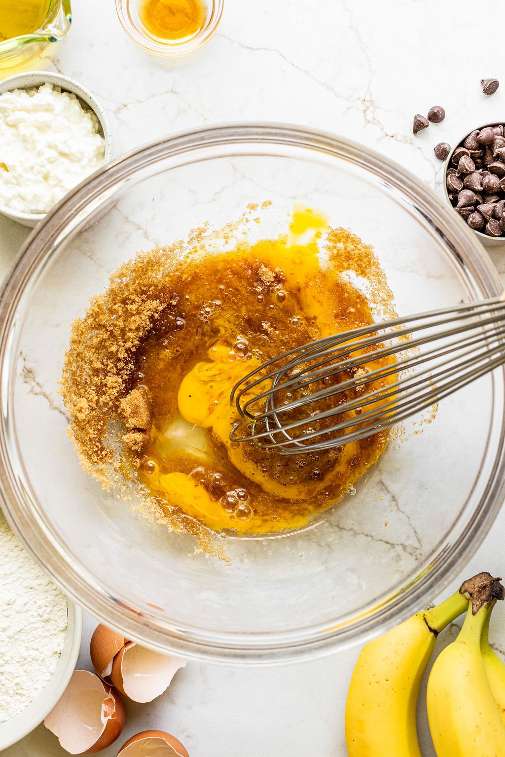 A whisk mixing eggs and brown sugar in a glass bowl, surrounded by bananas, chocolate chips, flour, cottage cheese, and broken eggshells on a white countertop.