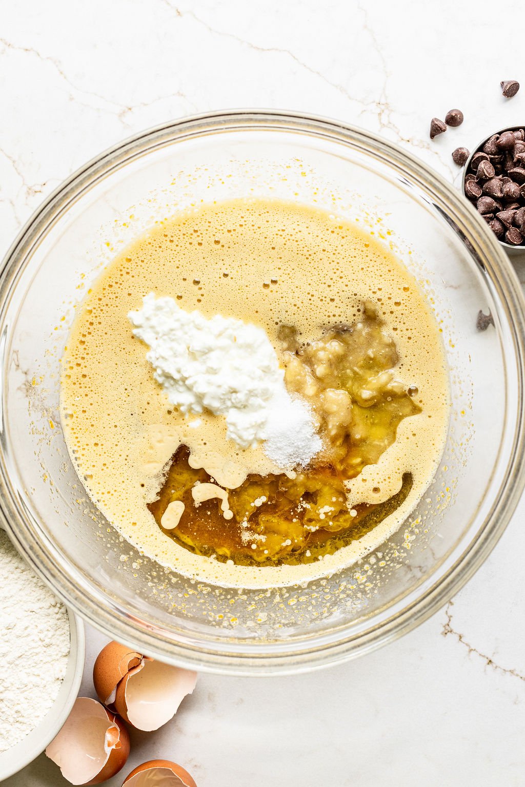 A glass bowl containing a mixture of beaten eggs, cottage cheese, mashed banana, and other baking ingredients.