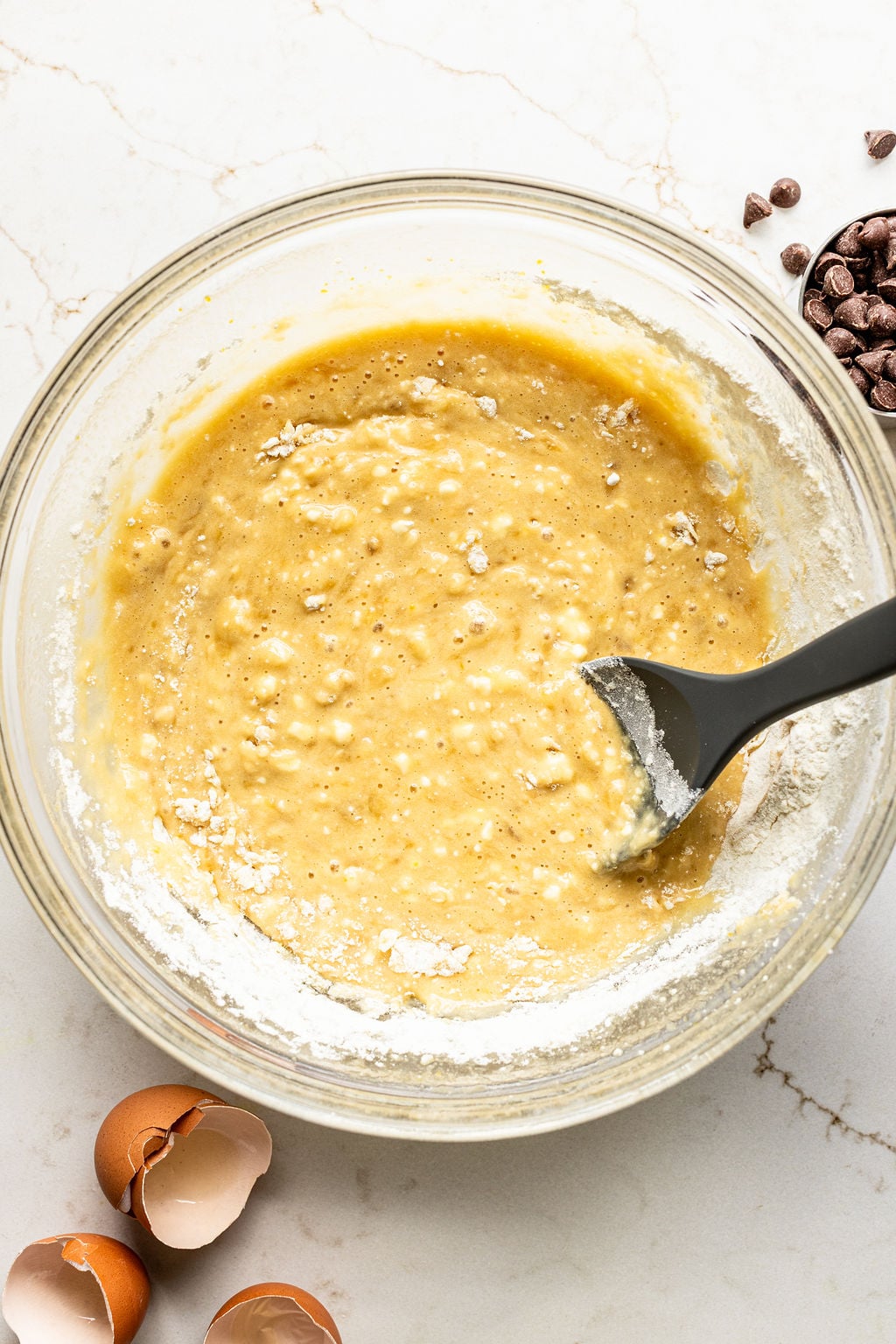 A glass bowl of banana bread batter is being mixed with a black spatula.