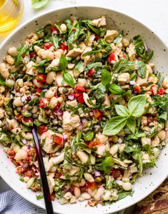 A bowl of mixed tuna salad with white beans, chopped red peppers, red onion, greens, herbs, and a black serving spoon. Slices of bread are nearby.