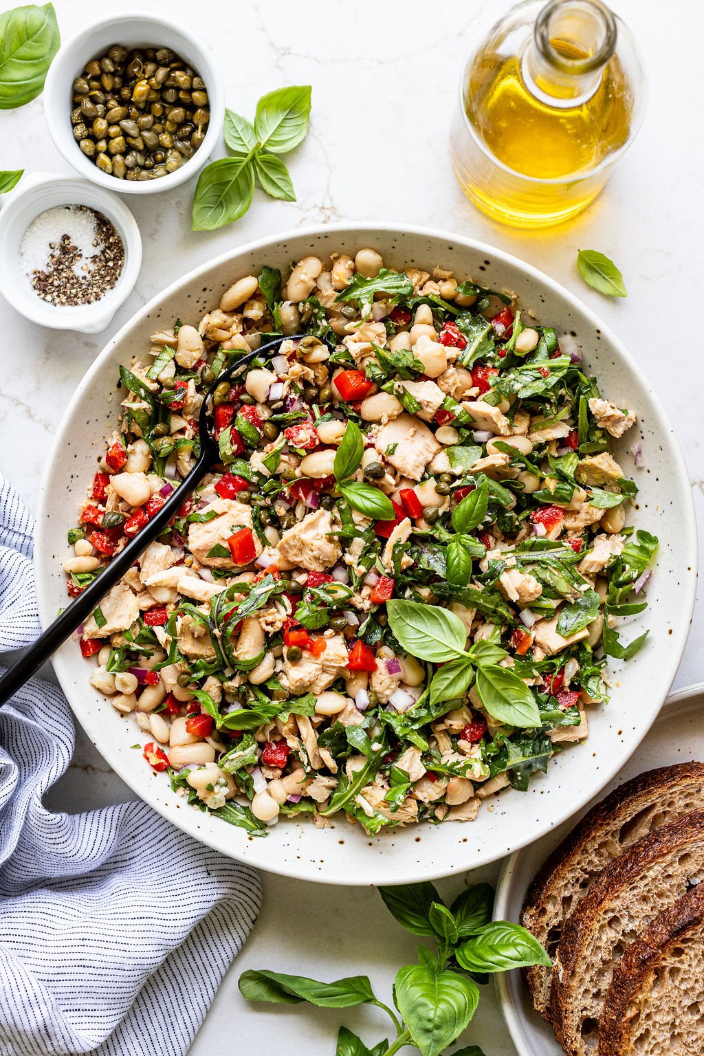 A large bowl of mixed salad with white beans, tuna, red peppers, basil, and greens, surrounded by a bottle of olive oil, capers, pepper, and slices of bread.