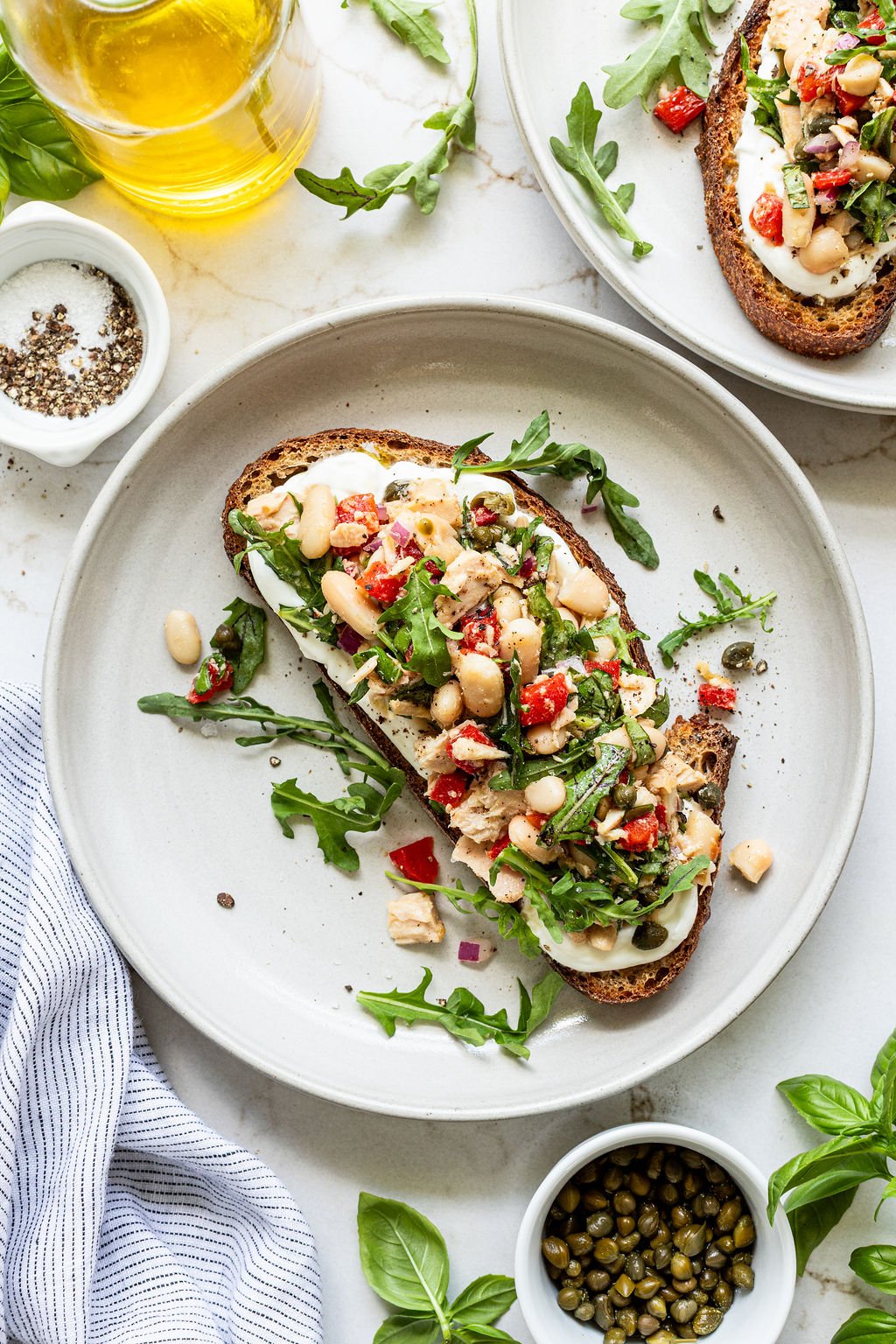 A slice of toasted bread topped with white beans, arugula, diced red peppers, capers, and herbs, served on a white plate.