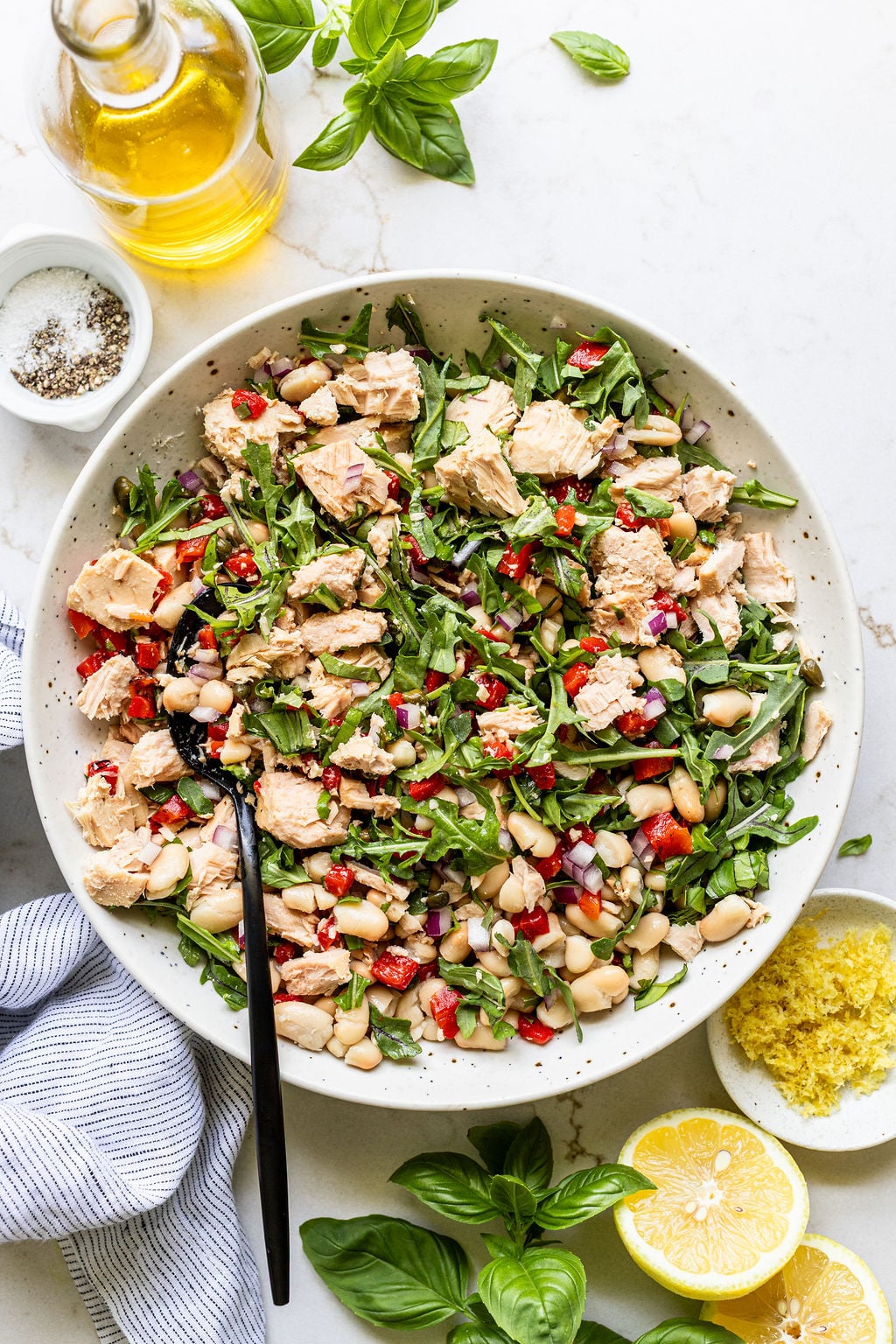 A bowl of tuna salad with white beans, chopped greens, red peppers, and onions, surrounded by lemon, basil, olive oil, and a jar of salt and pepper.