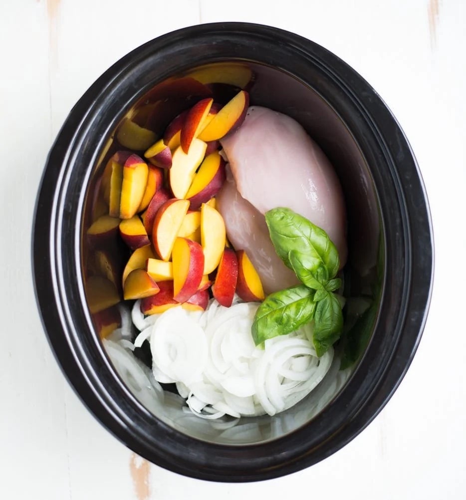 Sliced peaches, raw chicken breast, basil leaves, and sliced onions are arranged in a slow cooker.