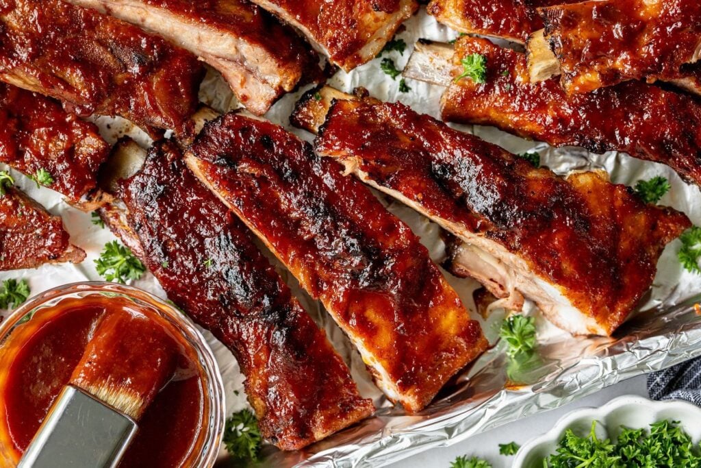 Barbecued pork ribs with a glossy sauce coating, garnished with parsley and served alongside a bowl of barbecue sauce and a basting brush.