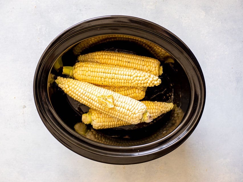 Four ears of corn inside a black slow cooker on a light-colored surface.