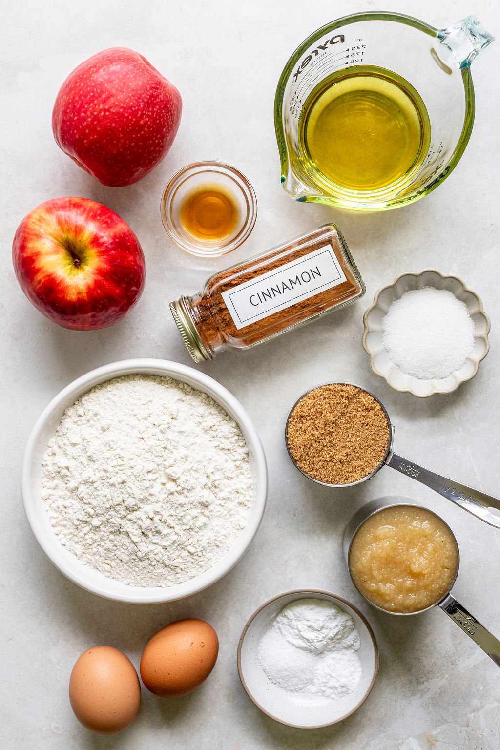 Top-down view of baking ingredients including apples, flour, eggs, oil, sugar, brown sugar, applesauce, cinnamon, baking powder, and vanilla extract arranged on a white surface.