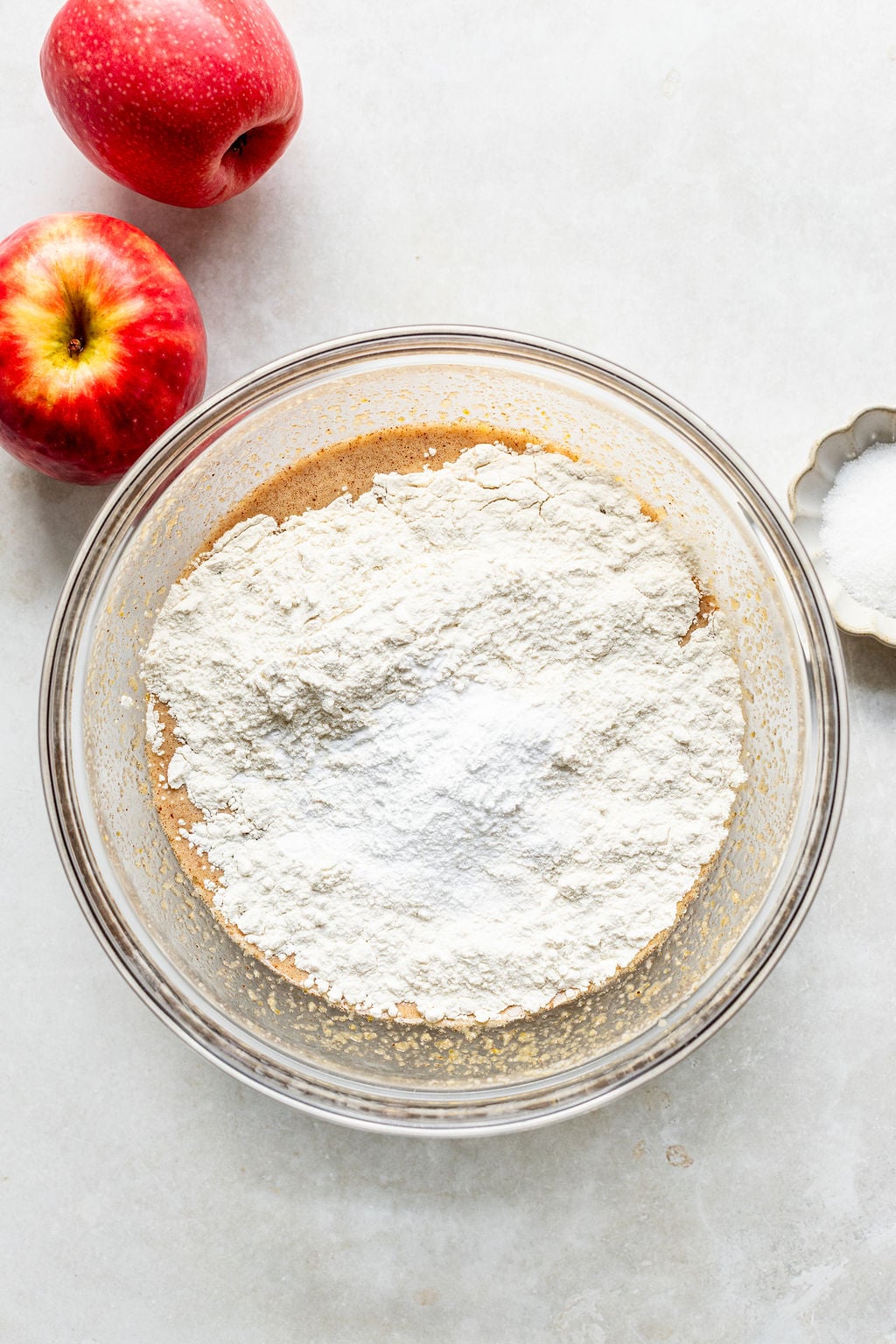 A glass bowl with flour on top of a brown batter mixture, next to two red apples and a small dish of sugar on a white surface.