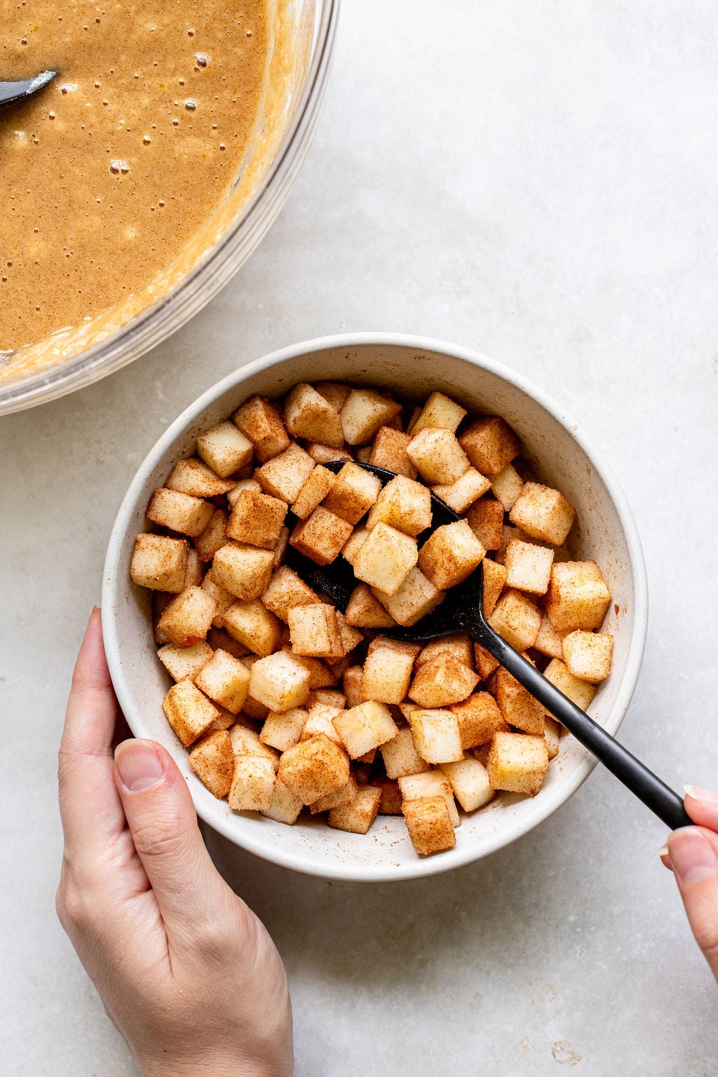 A hand holds a bowl of diced apples mixed with cinnamon, with another hand stirring them using a black spoon.