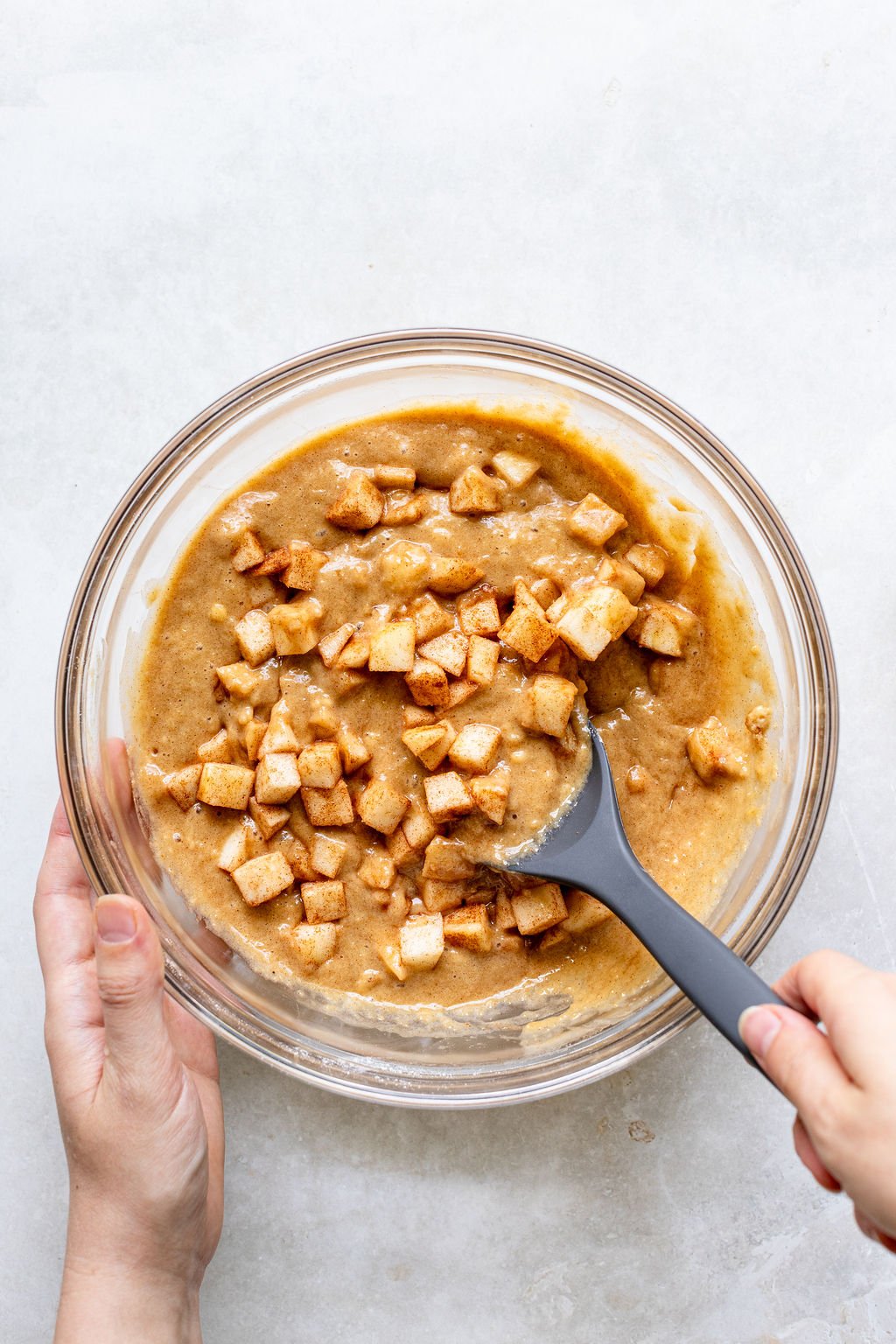 A person stirring a bowl of batter with chopped apples using a black spatula on a light-colored surface.