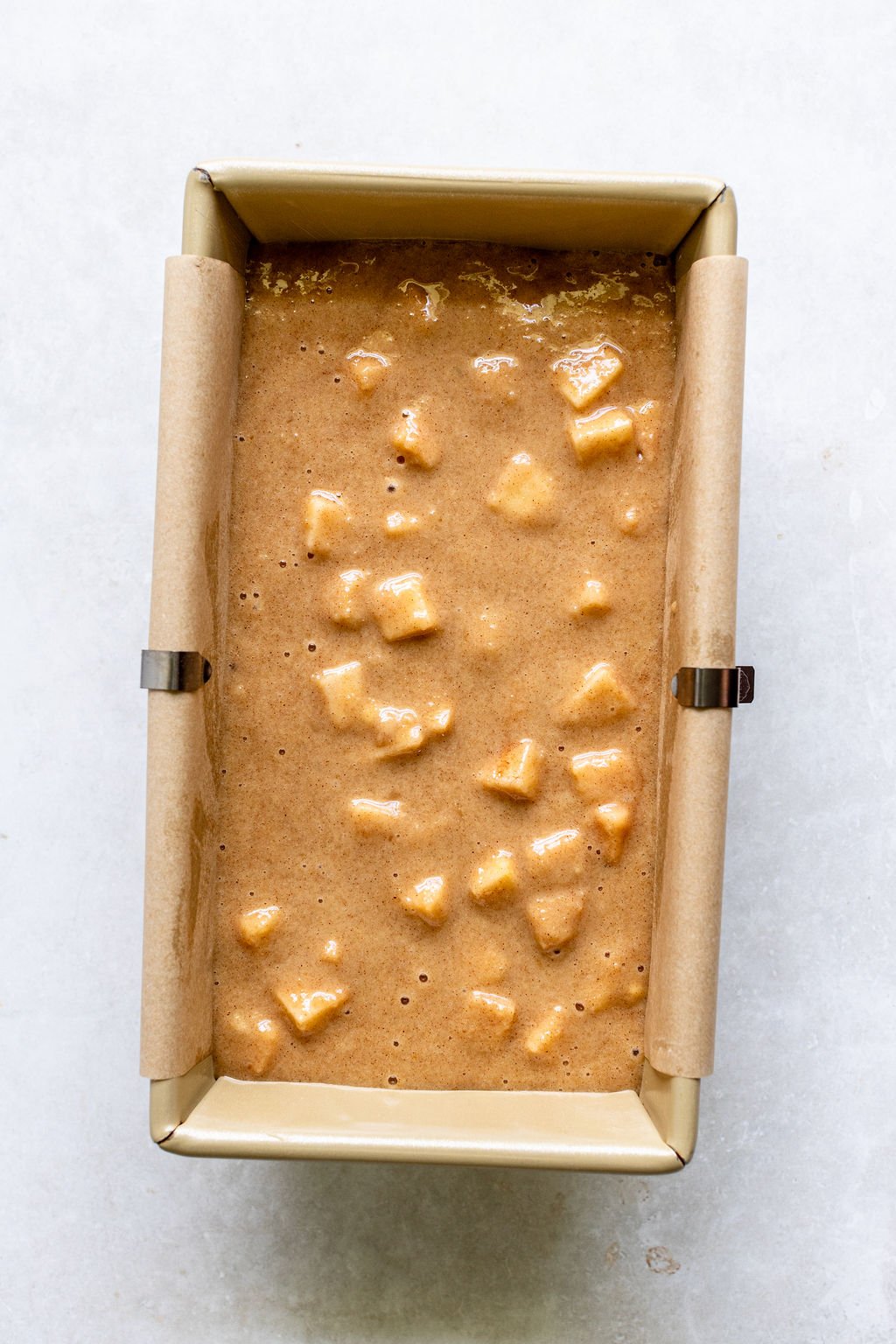 A parchment-lined loaf pan filled with light brown batter containing chunks of apples on a light-colored surface.