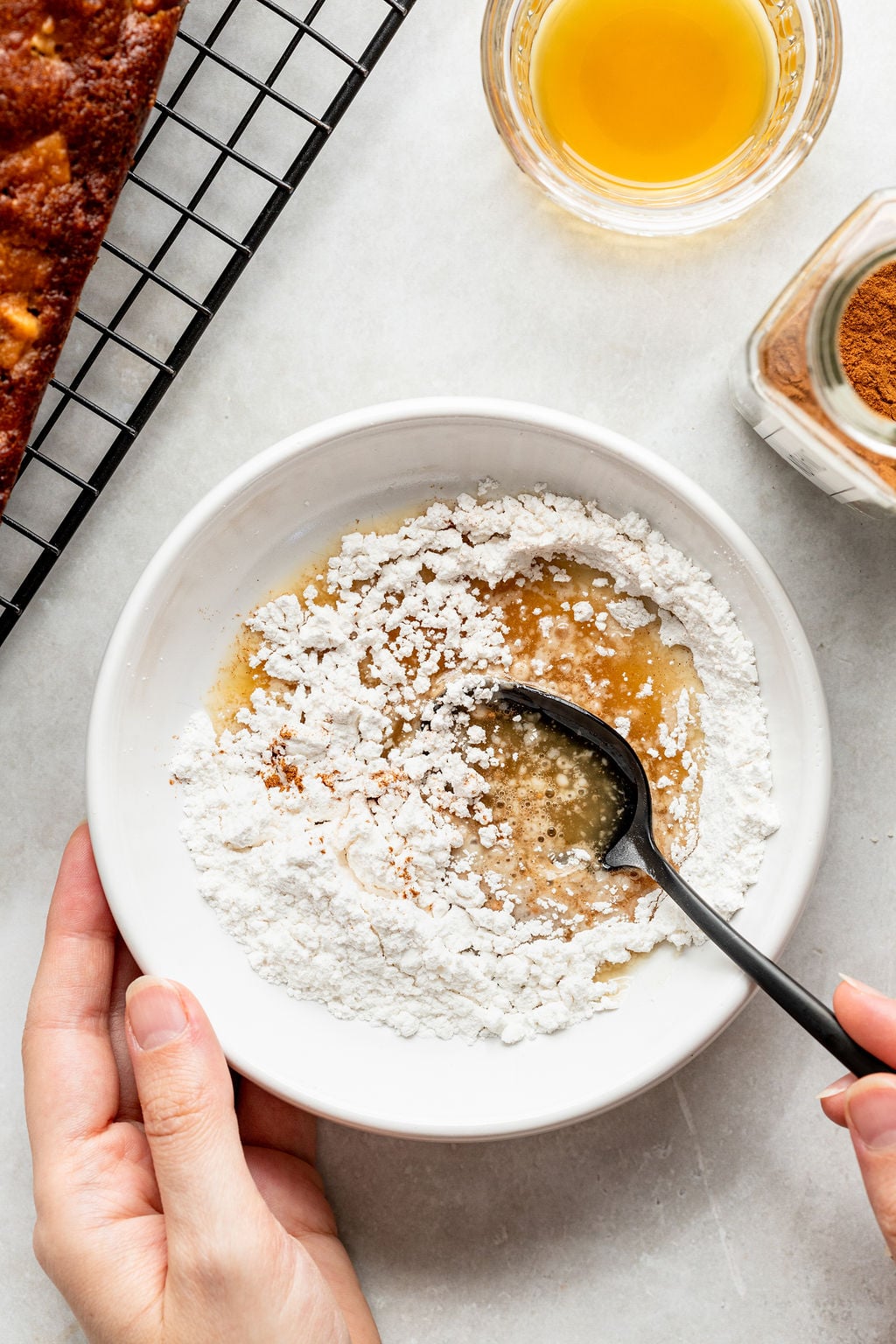 A hand holds a bowl with powdered sugar and liquid being mixed with a spoon.