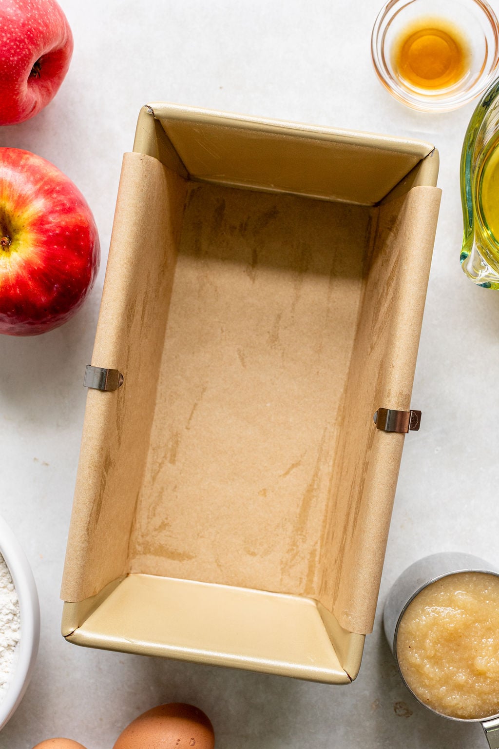 A parchment-lined loaf pan surrounded by apples, eggs, apple sauce, flour, oil, and vanilla on a light surface.