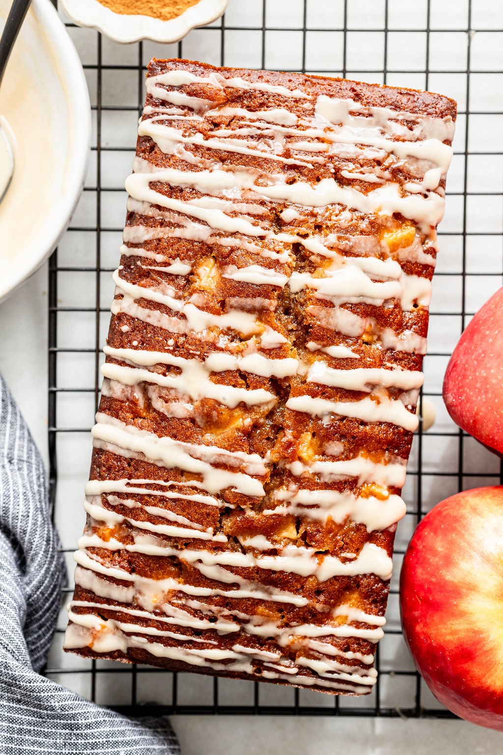 A glazed loaf of apple bread sits on a cooling rack next to two red apples.