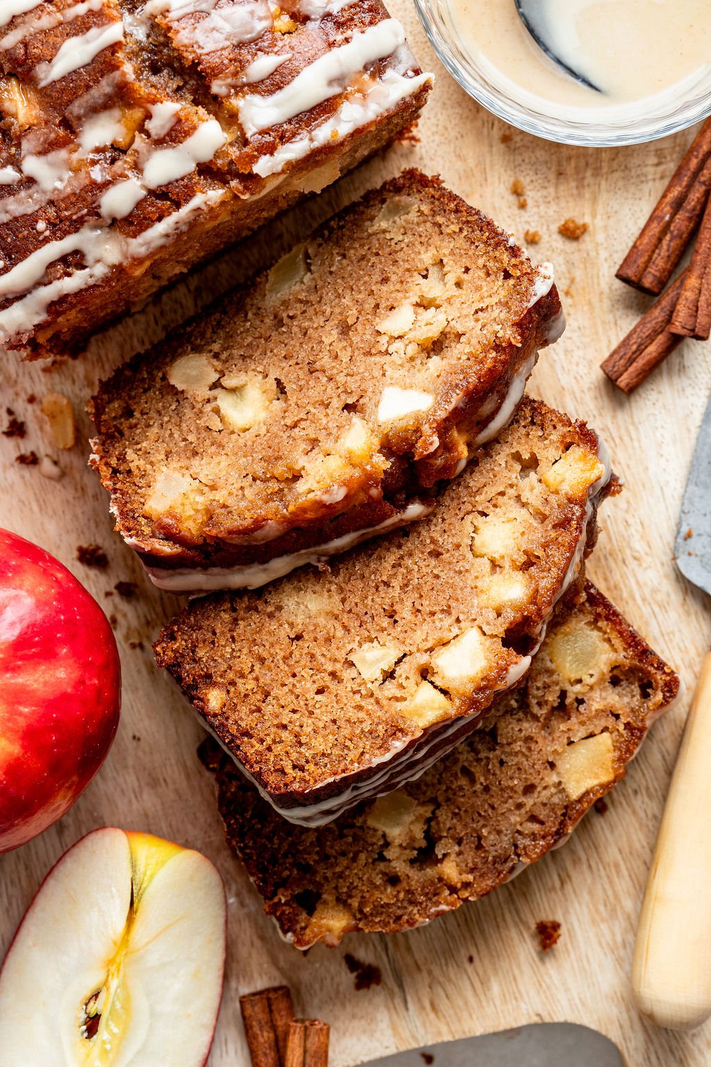 Sliced apple cinnamon loaf with icing on a wooden board, surrounded by a red apple, apple slices, cinnamon sticks, and a bowl of glaze.