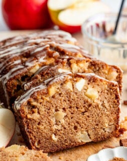 A sliced loaf of apple bread drizzled with icing, with visible apple chunks, sits on a wooden board near fresh apples and a small dish of cinnamon.