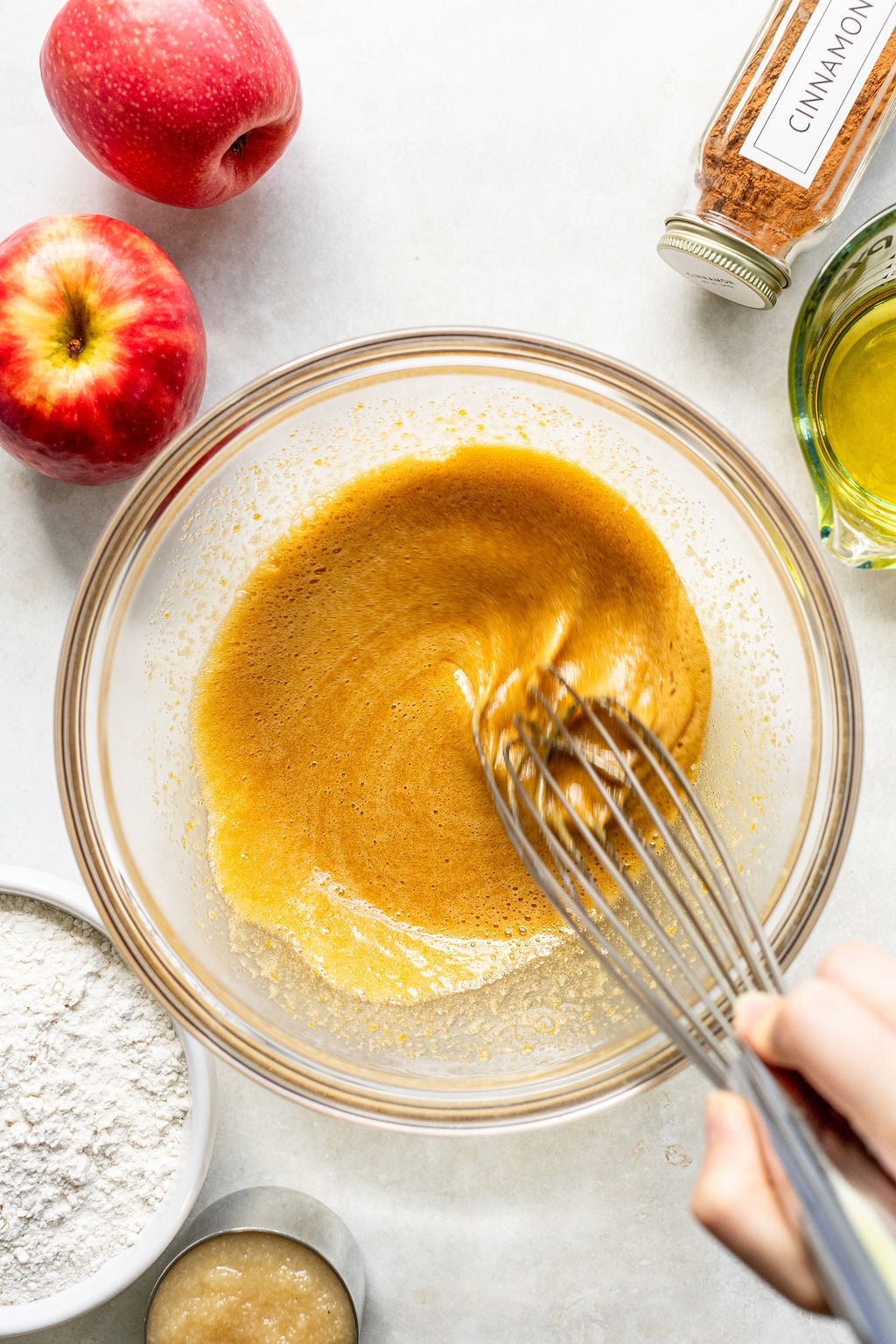 A hand whisking a brown batter in a glass bowl, surrounded by apples, flour, applesauce, cinnamon, and oil on a white surface.