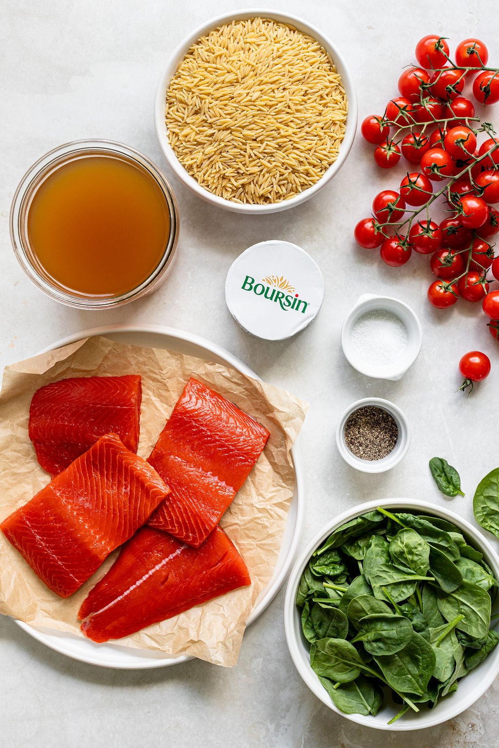 Top-down view of raw salmon fillets, orzo pasta, cherry tomatoes, spinach, broth, Boursin cheese, salt, and pepper arranged on a light surface.