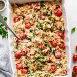 A white baking dish filled with baked orzo pasta, cherry tomatoes, spinach, and herbs, with a serving spoon and garnished with fresh parsley.