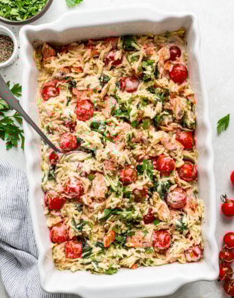 A white baking dish filled with baked orzo pasta, cherry tomatoes, spinach, and herbs, with a serving spoon and garnished with fresh parsley.
