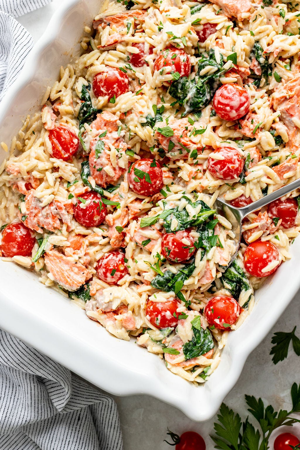 Baked orzo pasta with cherry tomatoes, spinach, shredded carrots, and a creamy sauce, served in a white baking dish with a serving spoon.