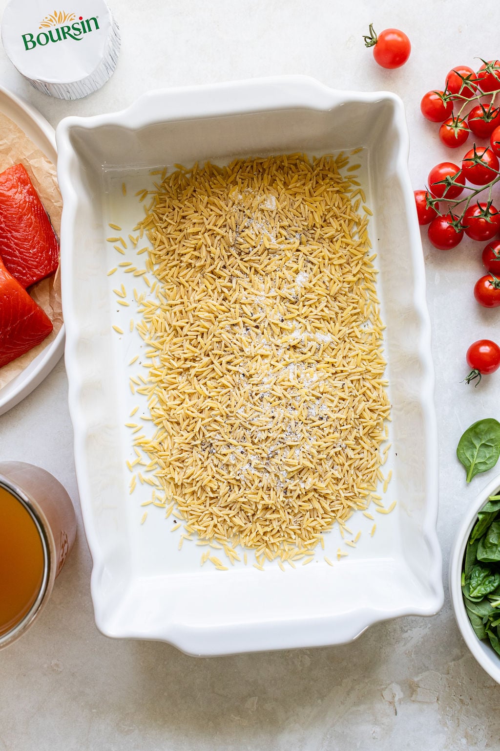 A white baking dish filled with uncooked orzo pasta, surrounded by salmon, cherry tomatoes, spinach, broth, and a package of Boursin cheese.