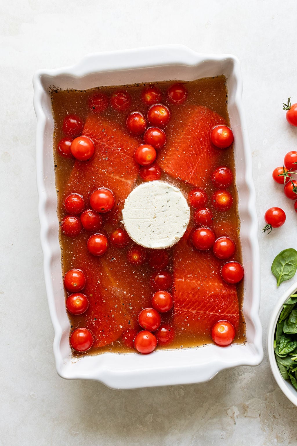 White baking dish with raw salmon fillets, cherry tomatoes, and a round block of cheese in the center, all in a brown liquid.