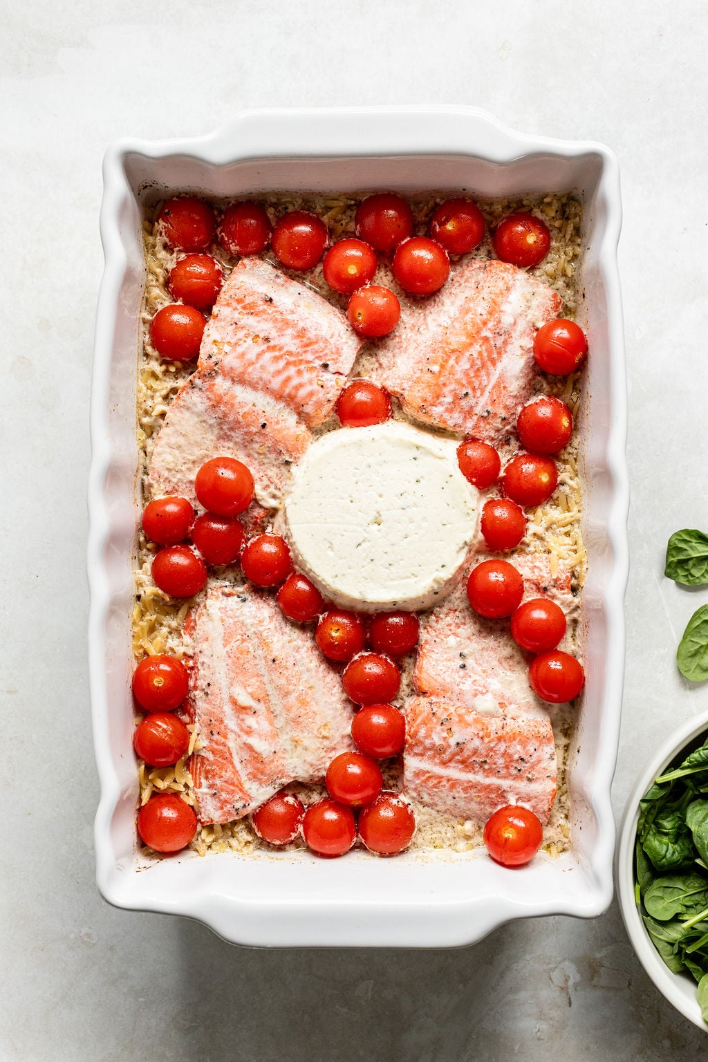 Uncooked salmon fillets, cherry tomatoes, and a round of cheese are arranged on top of pasta in a white baking dish.