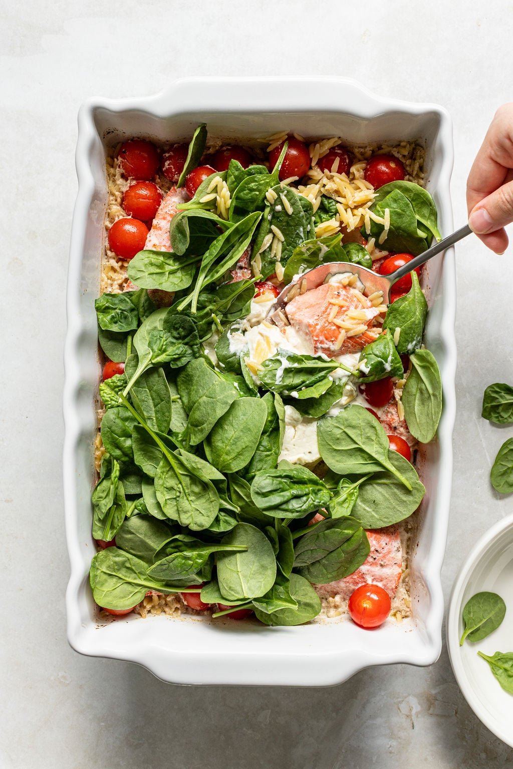 A hand holding a spoon is scooping a portion from a baking dish filled with spinach, cherry tomatoes, orzo pasta, and creamy sauce.