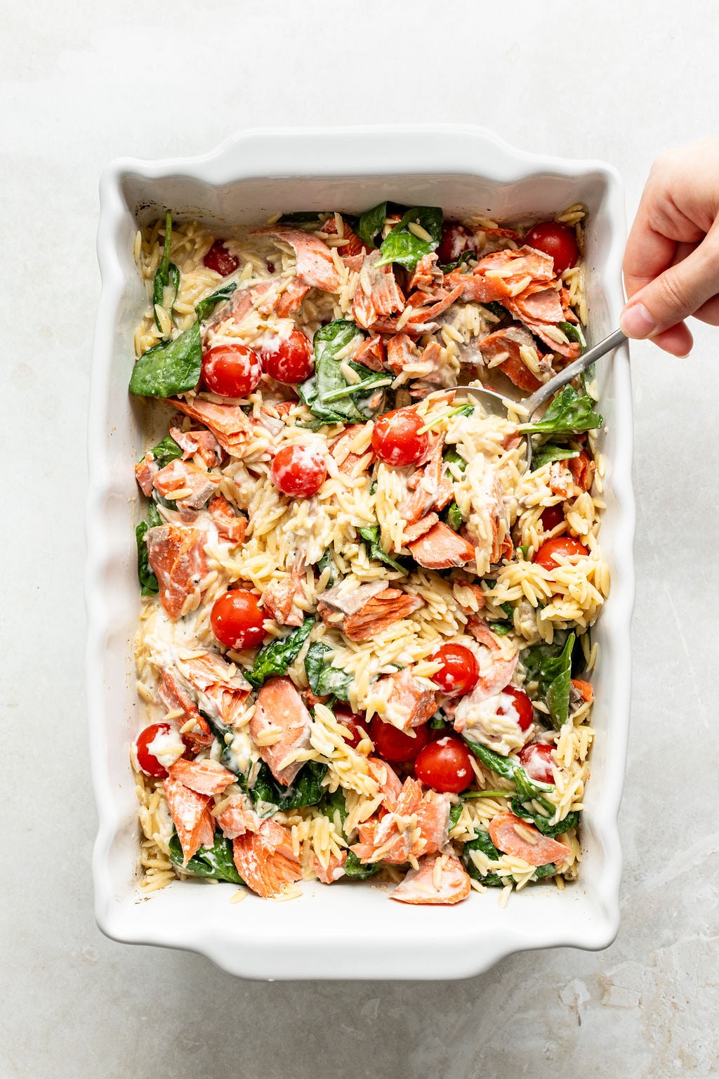 A white baking dish filled with orzo pasta, cherry tomatoes, spinach, flaked salmon, and creamy sauce. A hand holds a fork, mixing the ingredients.