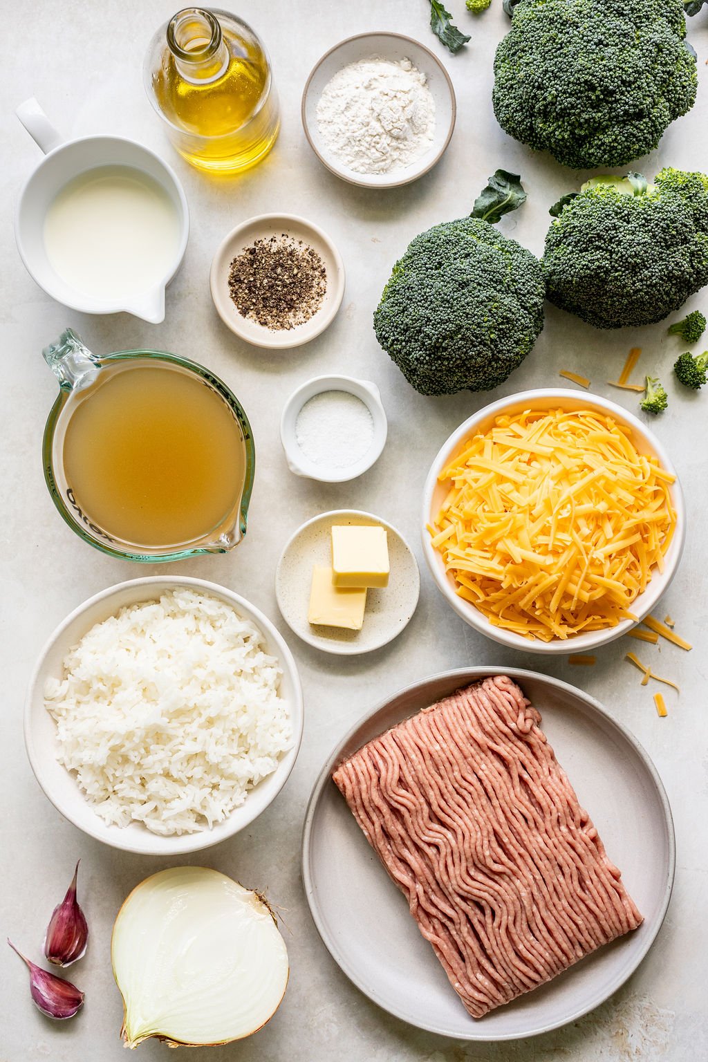 Ingredients for a broccoli cheese casserole arranged on a counter, including broccoli, shredded cheese, ground meat, cooked rice, broth, butter, onion, garlic, flour, pepper, and salt.