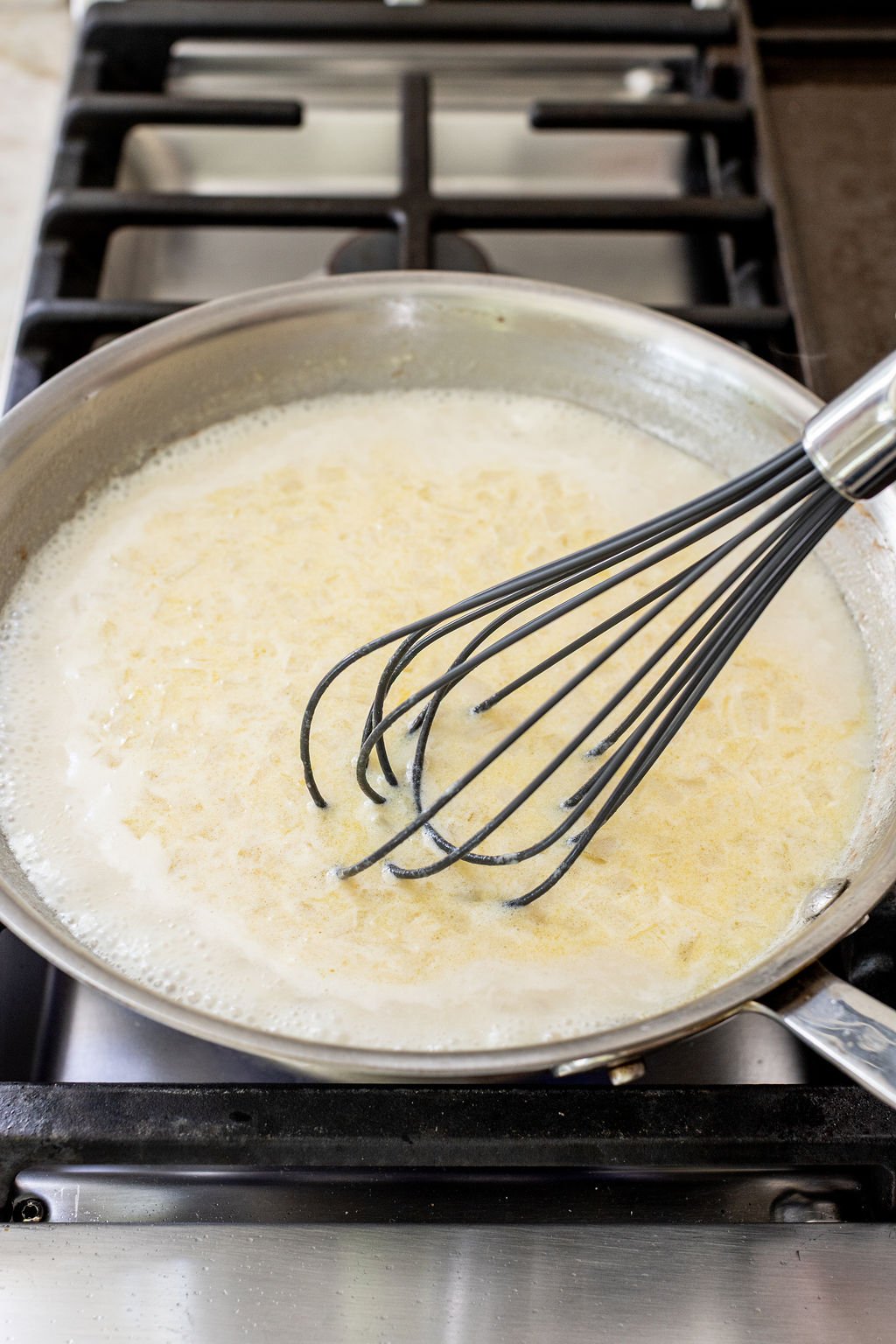 A whisk stirring a creamy sauce with grated cheese in a stainless steel pan on a gas stove.