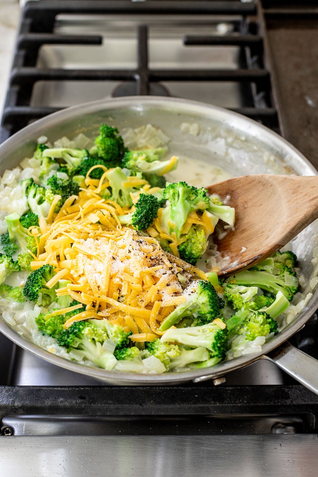 Broccoli, shredded cheddar cheese, and onions in a pan being stirred with a wooden spoon on a stovetop.